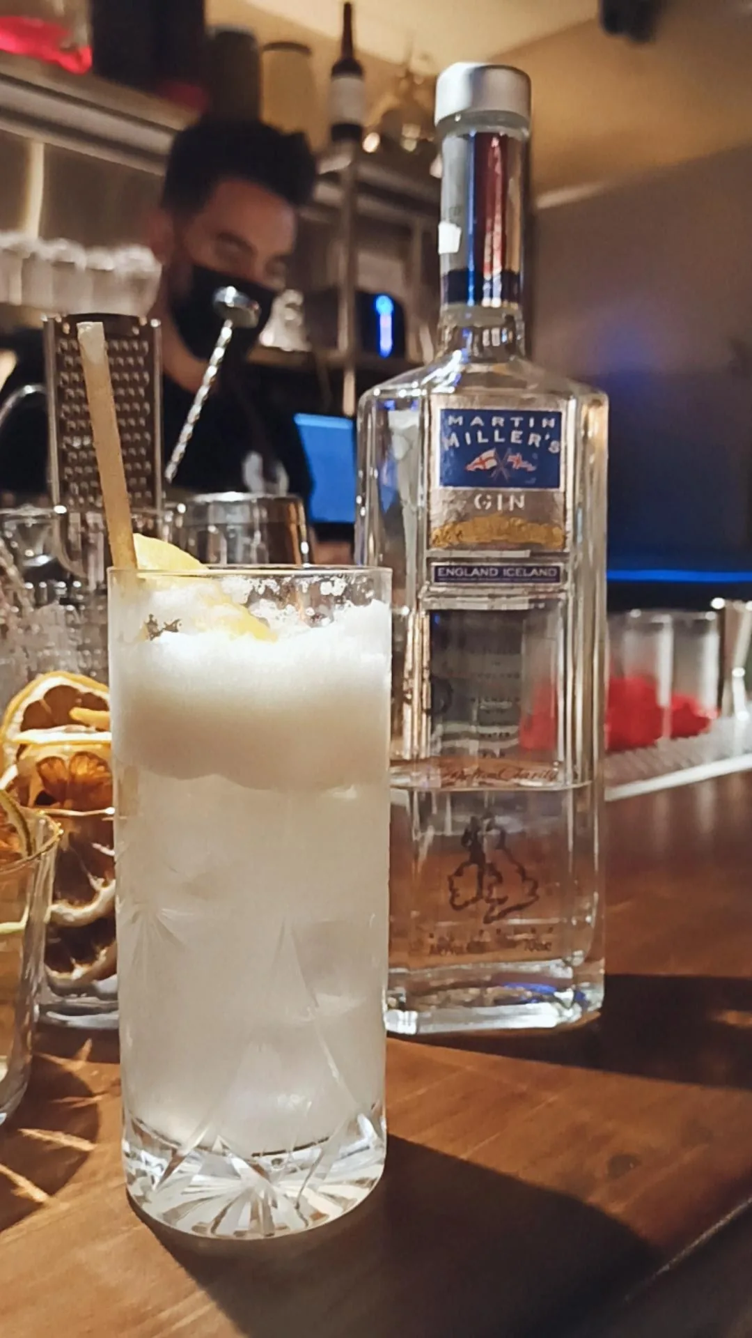 A tall cocktail in a glass with ice beside a vodka bottle on a wooden bar