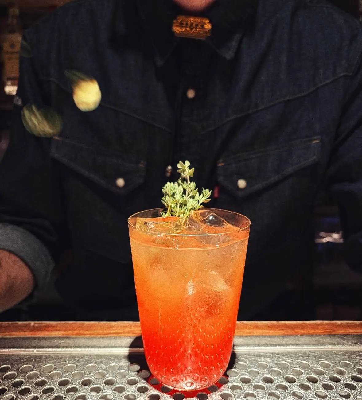 An orange-hued cocktail garnished with a sprig of herb on a dark bar counter