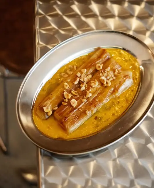 Grilled leeks in a curried yellow sauce topped with chopped hazelnuts on an oval metal platter