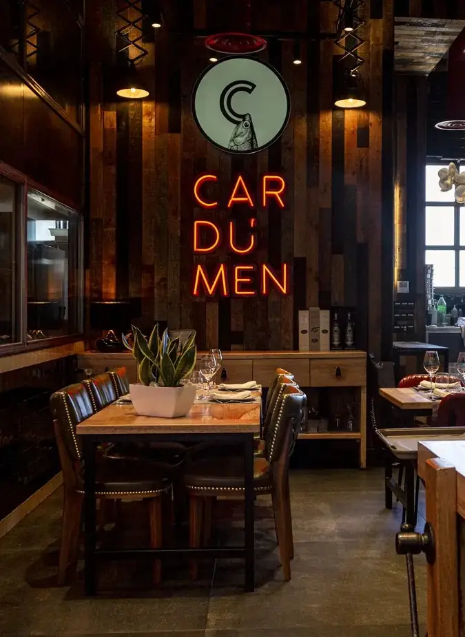 Dining room with reclaimed-wood walls, a set table with wine glasses and a plant centrepiece beneath the venue's fish logo and orange CARDUMEN neon sign