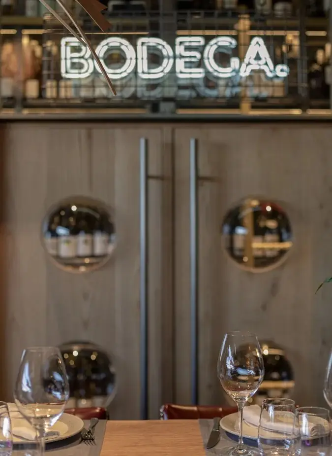 Table detail with wine glasses and place settings, a white BODEGA neon sign and wine bottles behind porthole windows in the background