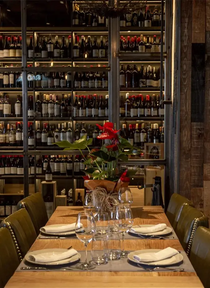 Set wooden dining table with wine glasses and a red flower centrepiece in front of a floor-to-ceiling backlit wine wall
