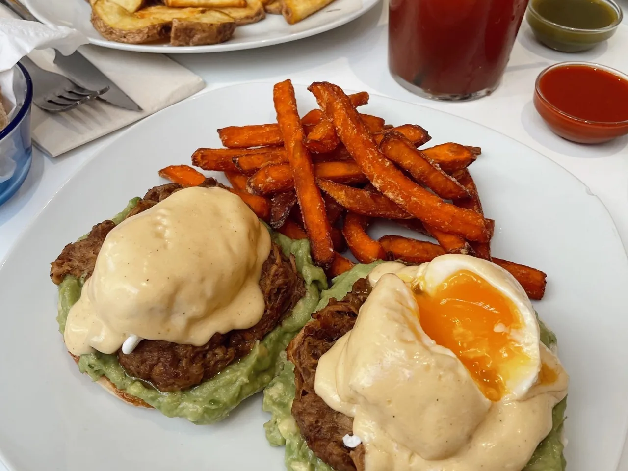 Two toasted rounds topped with avocado, poached eggs and hollandaise, served with sweet potato fries on a white plate