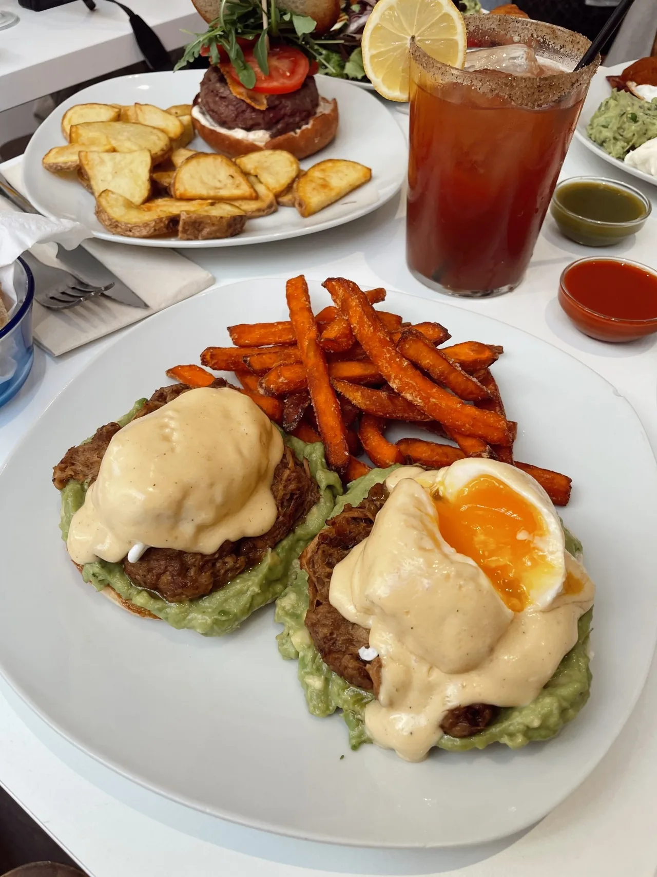 A plate of poached eggs with hollandaise on avocado toast with sweet potato fries, and a tall glass of red drink beside it