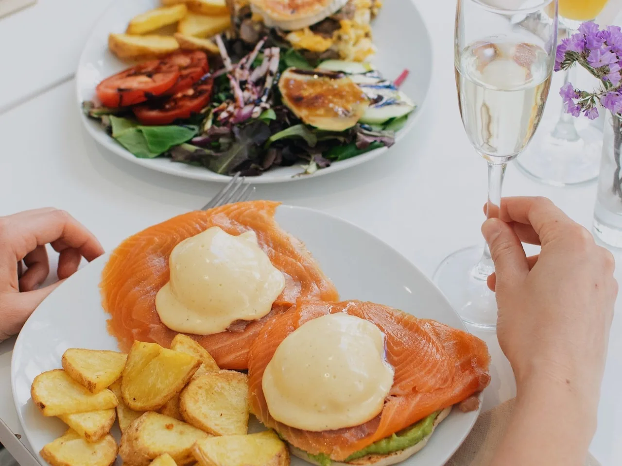 A plate of sliced salmon with poached eggs and hollandaise beside roasted potatoes, with a glass of sparkling wine