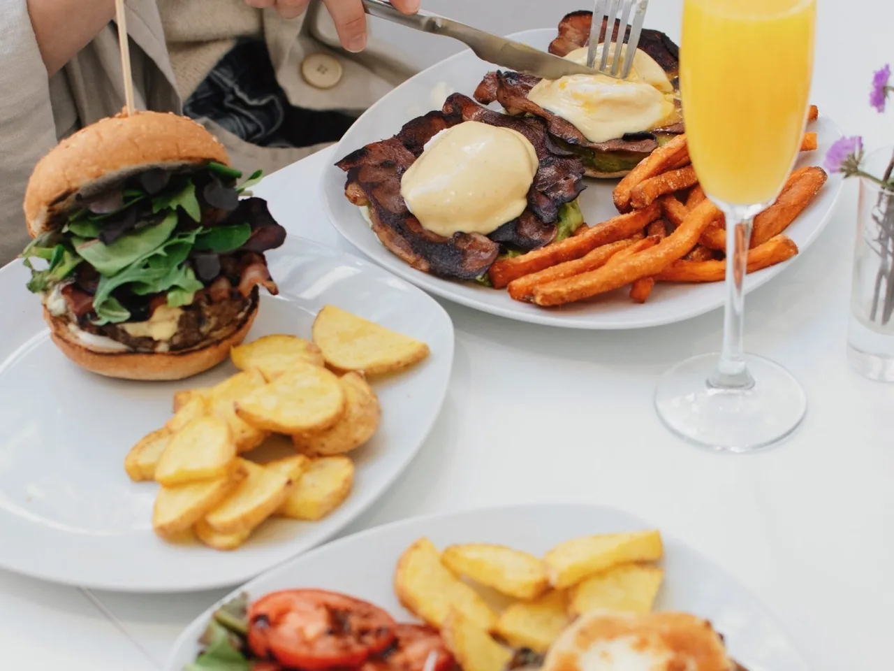 A brunch table with a burger, eggs with hollandaise, sweet potato fries, roasted potatoes and a glass of orange juice