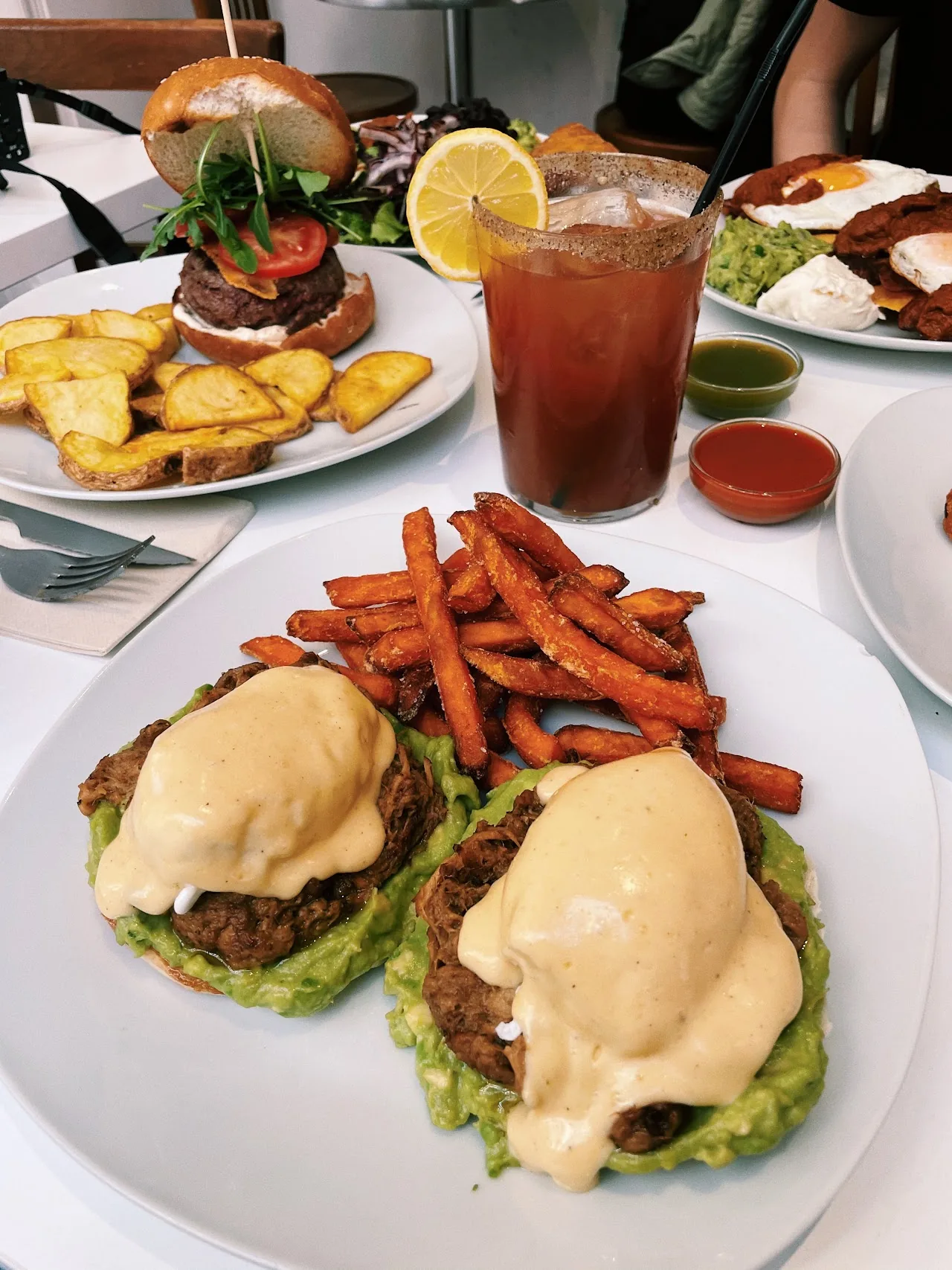 Two eggs Benedict on avocado toast beside sweet potato fries and a Bloody Mary