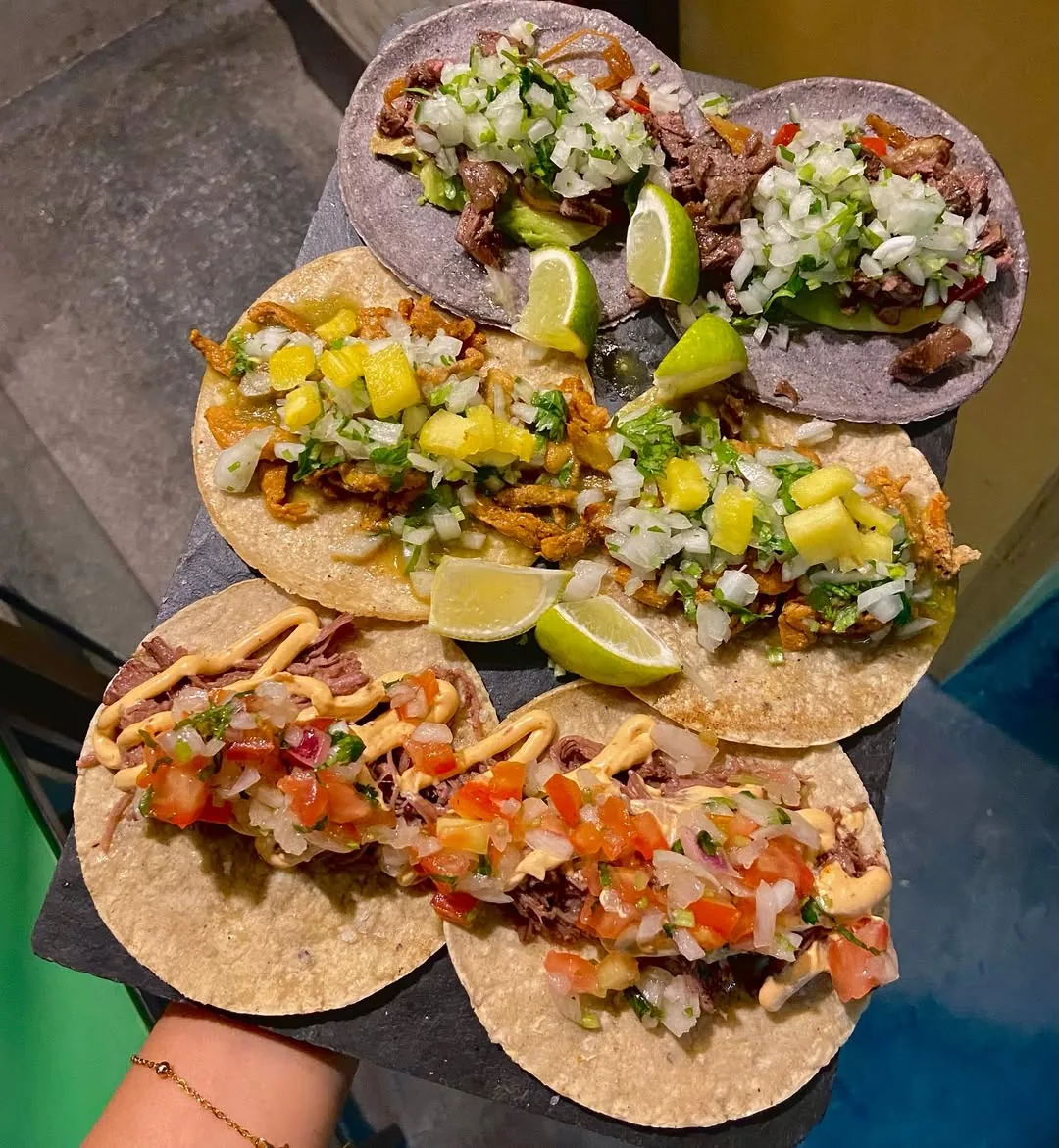 Five loaded street tacos on corn and flour tortillas, topped with pulled meat, onion, cilantro, pineapple, and lime wedges, held on a wooden board