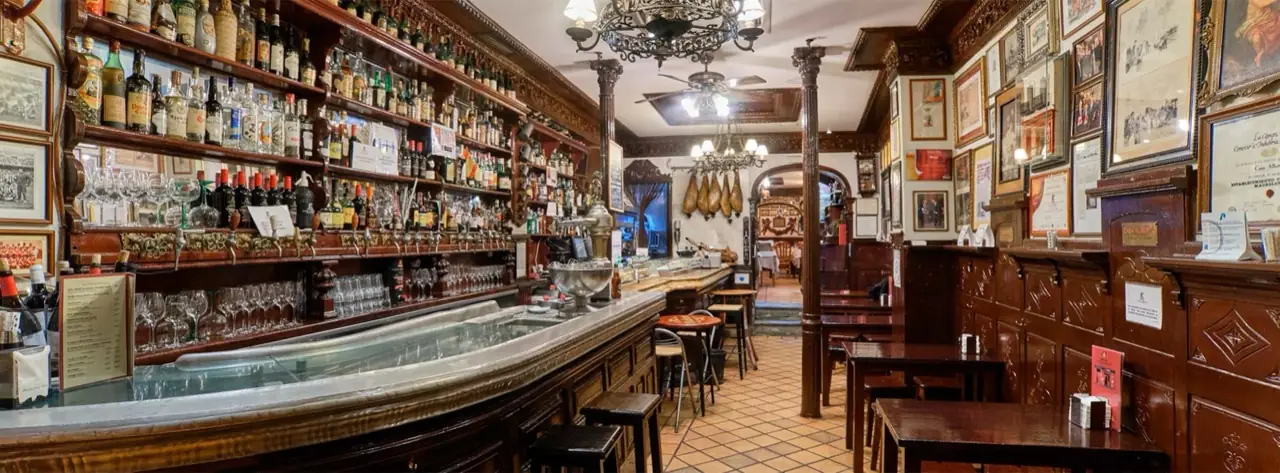Long carved mahogany bar of a traditional Madrid tavern backed by shelves crowded with liquor bottles, hanging jamon legs, chandeliers and framed photographs