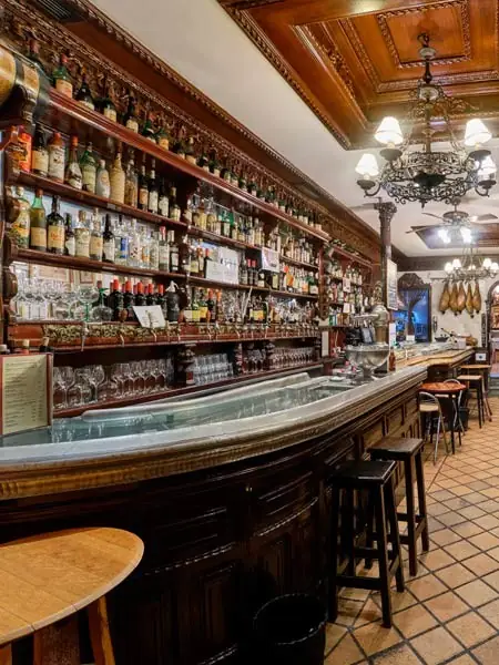 Curved wooden tavern bar with shelves of bottles, ornate chandelier, hanging jamon and stools on a tiled floor