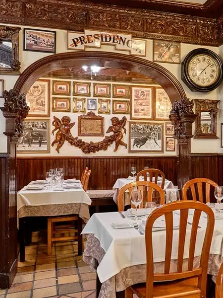 Dining room with white-clothed tables and wooden chairs beneath an arch of framed bullfighting prints, carved cherubs and a wall clock
