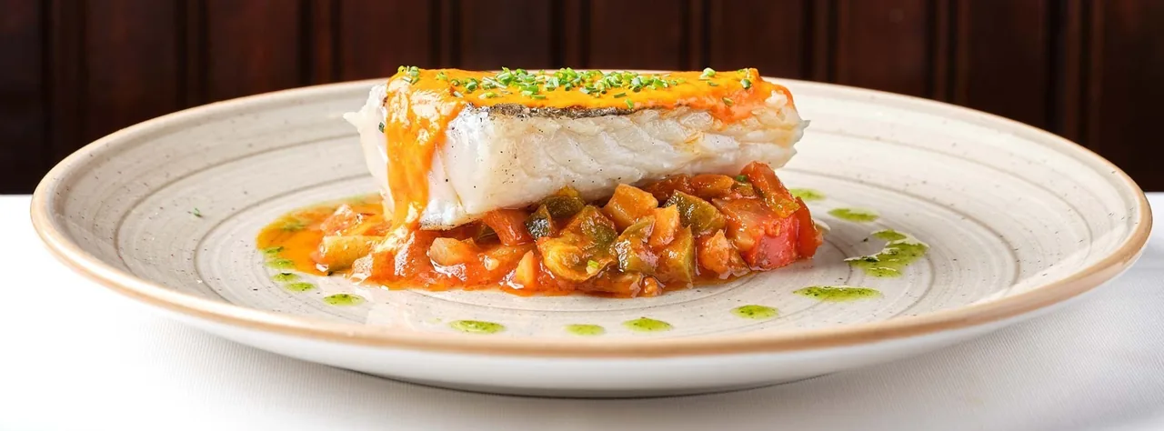 A thick piece of white fish topped with orange sauce on a bed of diced vegetables on a round plate