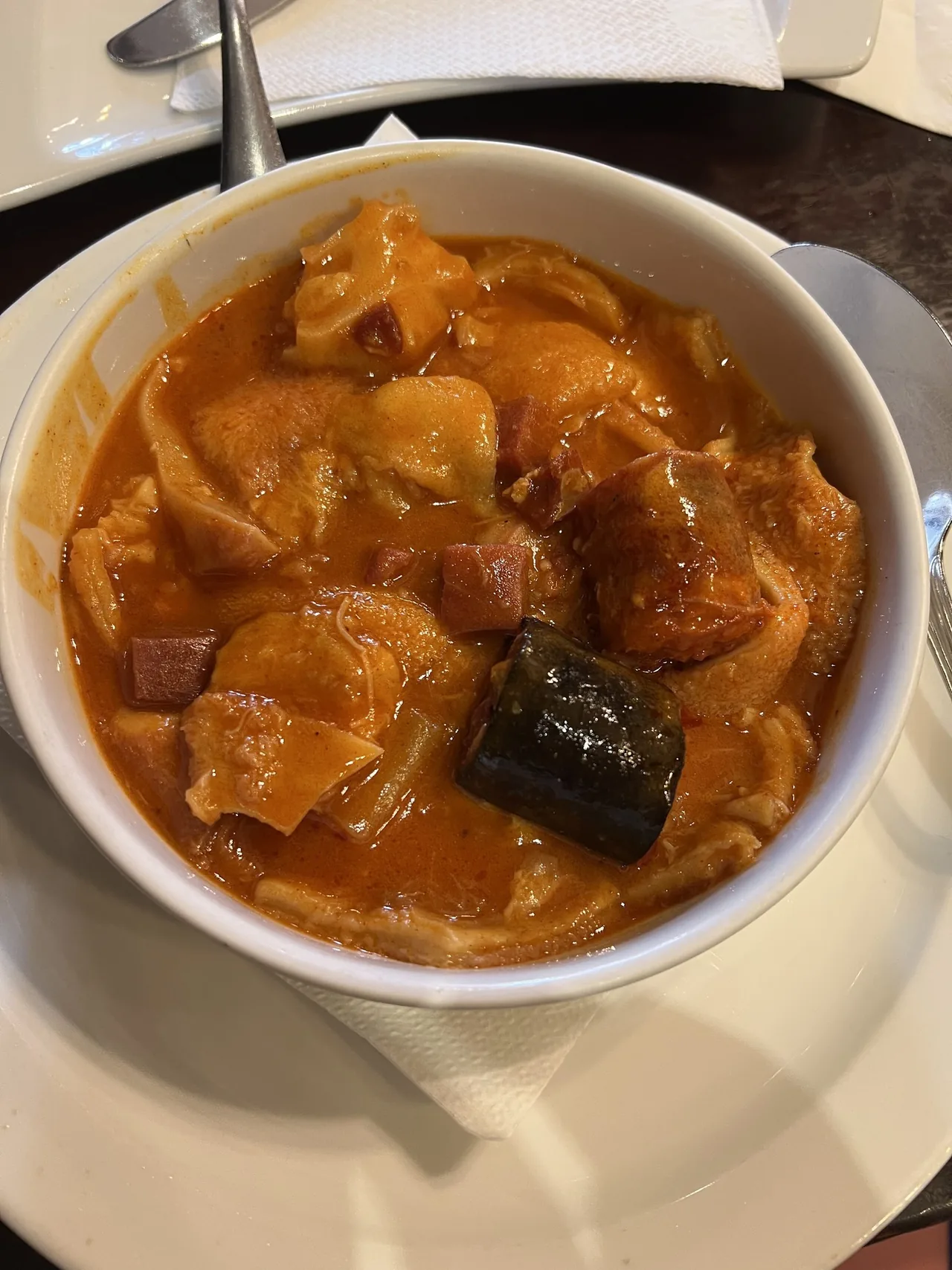 A white bowl of tripe stew in orange sauce with chorizo pieces and a slice of black pudding