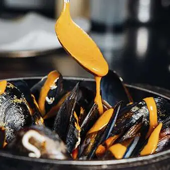 Mussels in a black pot topped with a golden-orange sauce being drizzled from a spoon, Casa Amàlia 1950 Port Vell