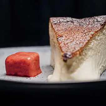 Slice of baked cheesecake dusted with powdered sugar next to a small red fruit compote cube on a dark plate, Casa Amàlia 1950 Port Vell