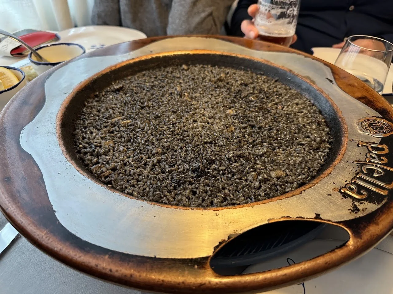 Black rice in a shallow paella dish set in a wide ceramic holder