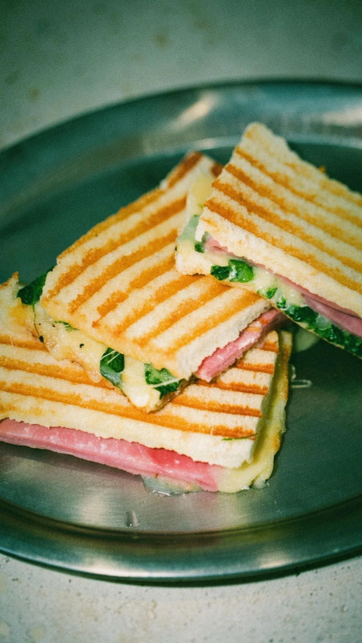 Grill-marked toasted sandwiches with ham, cheese and green leaves stacked on a metal plate