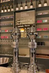 Bar interior with twin chrome beer towers, a chalkboard menu and shelves of wine bottles behind the counter