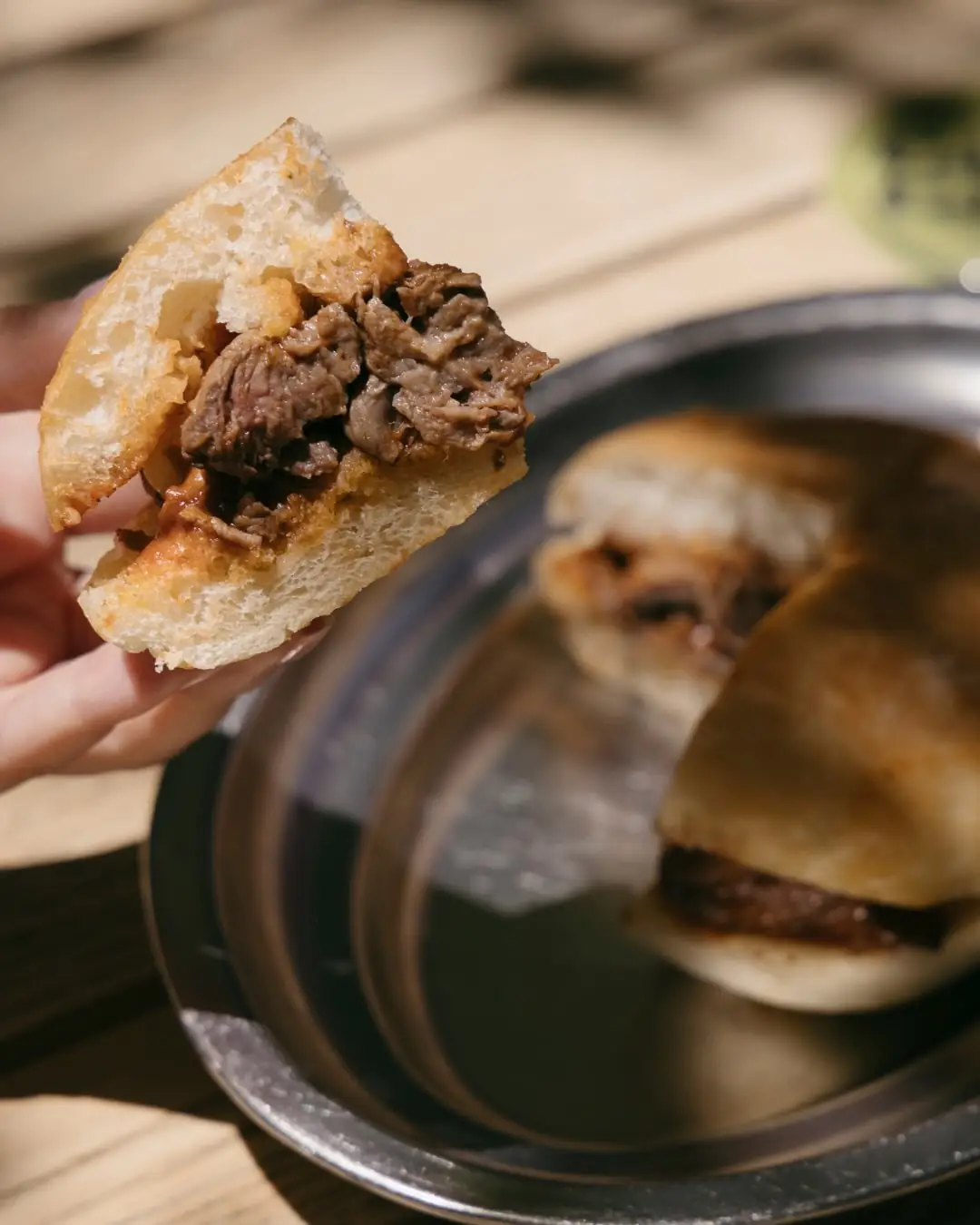 Hand holding a torn beef sandwich showing shredded meat filling, with two more sandwiches on a plate behind