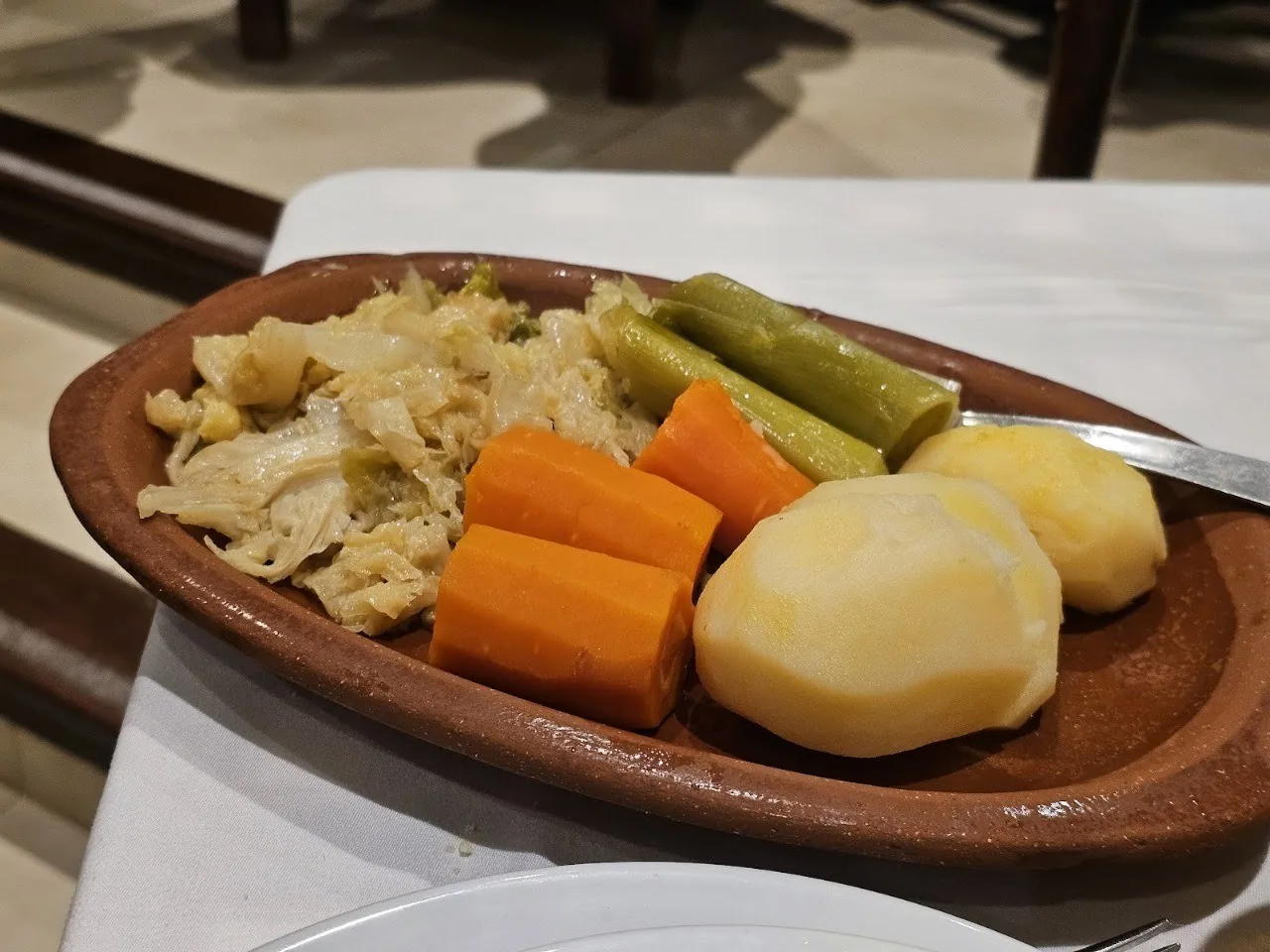 An oval terracotta dish of boiled cabbage, leeks, carrots and potatoes on a white tablecloth