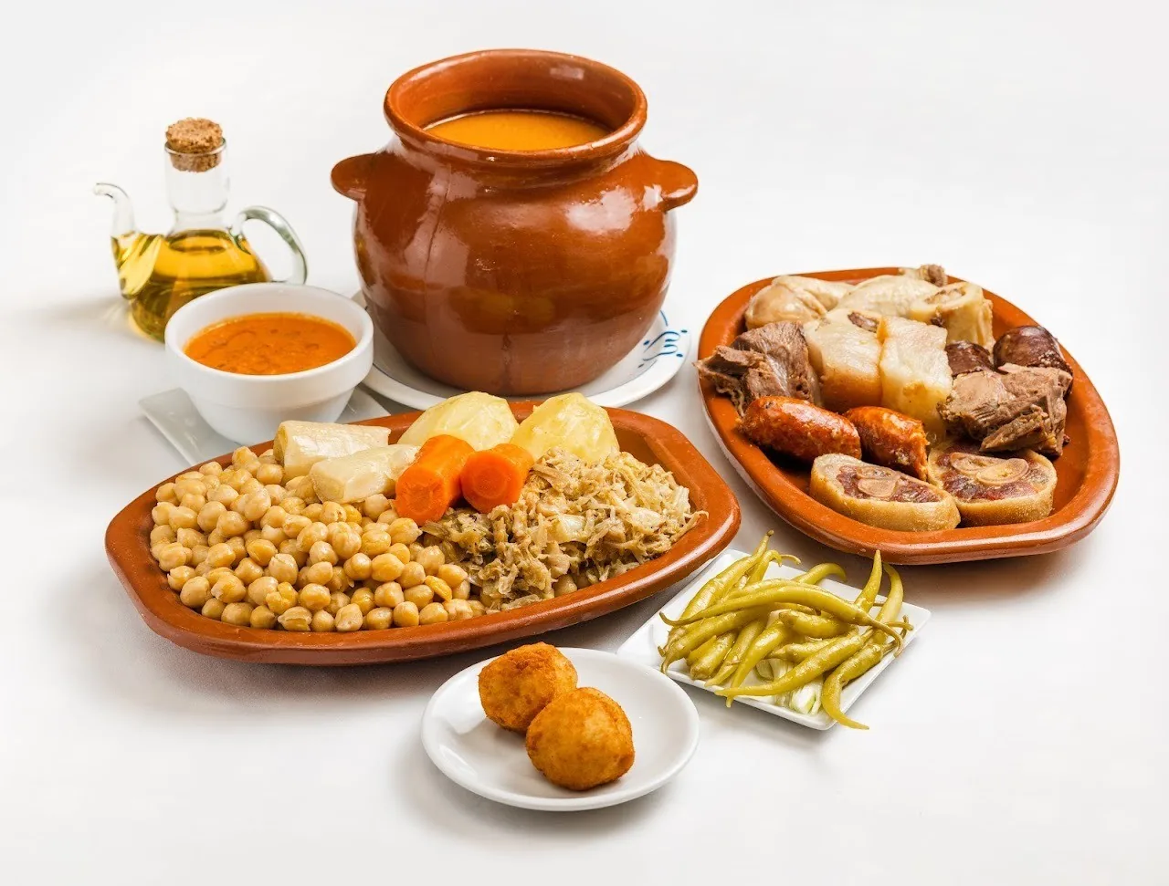 A clay pot of broth, a platter of chickpeas, vegetables and meats, and a side of fried potatoes on a white background