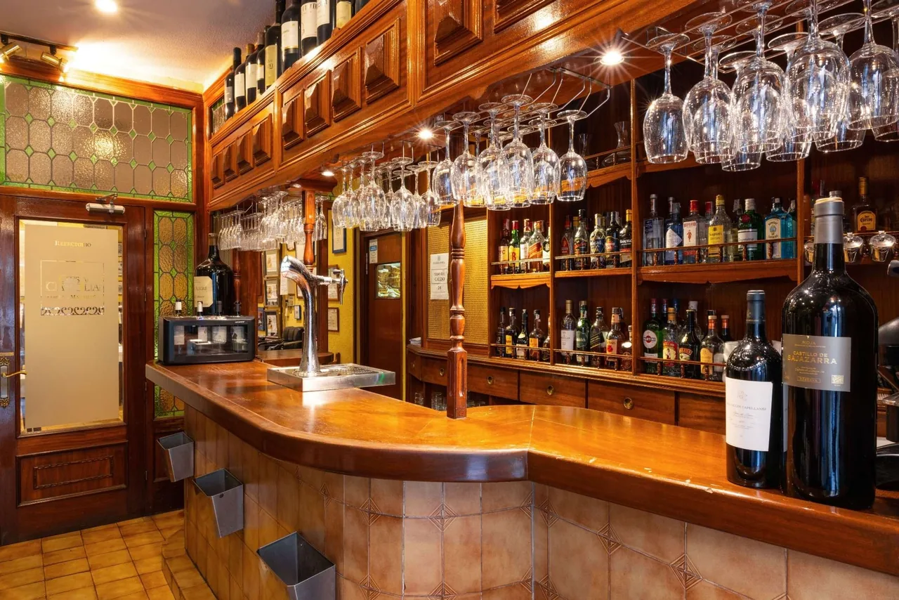 A wooden bar counter with shelves of bottles and hanging glasses