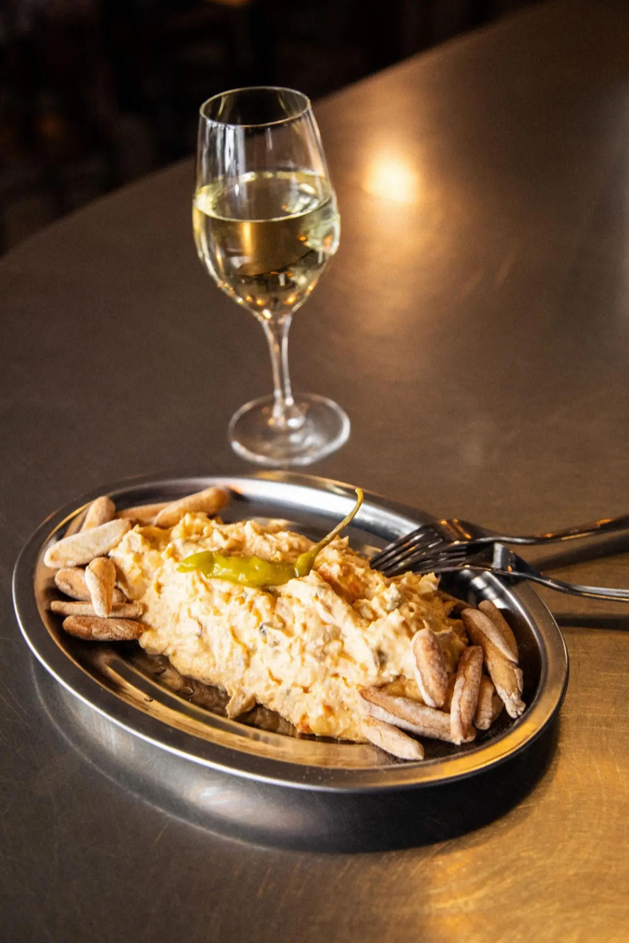 Oval metal plate of creamy ensaladilla topped with a pickled green pepper, ringed with breadsticks, beside a glass of white wine on a dark bar top