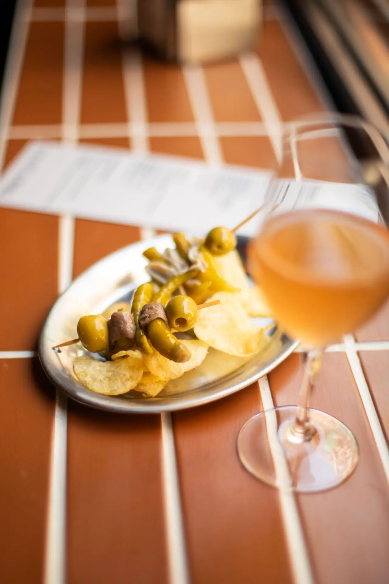 Metal plate of gilda skewers of green olives, anchovy and pickled guindilla peppers over potato crisps, with an orange-hued vermouth glass on a terracotta-tiled table