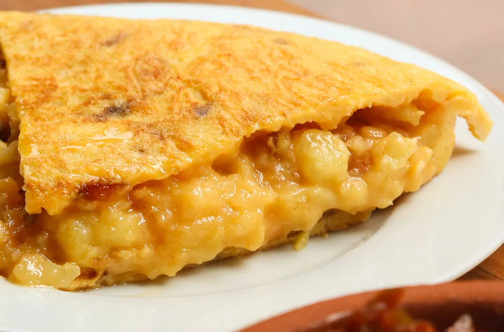A wedge of Spanish potato omelette on a white plate in tight close-up, the cut face showing loose barely-set potato and egg spilling out