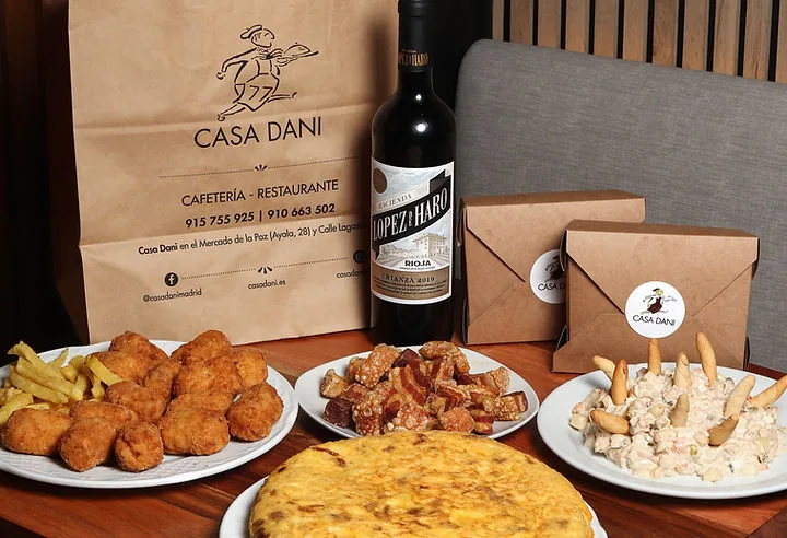 A takeaway spread on a wooden table, a plate of croquettes with chips, a plate of pork crackling, a plate of Russian salad with breadsticks, a whole potato omelette, a bottle of Rioja and takeaway boxes beside a printed Casa Dani paper bag