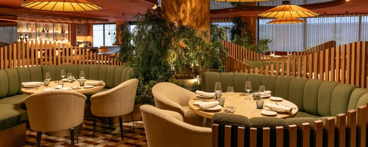Warmly lit dining room with green boucle banquettes, cream tub chairs, terrazzo tables, wooden slat dividers, hanging pleated lamps and hanging greenery