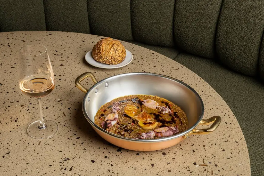 Copper pan of soupy rice with squid and roasted onion drizzled with dark sauce, beside a glass of rose wine and a seeded bread roll on a terrazzo table