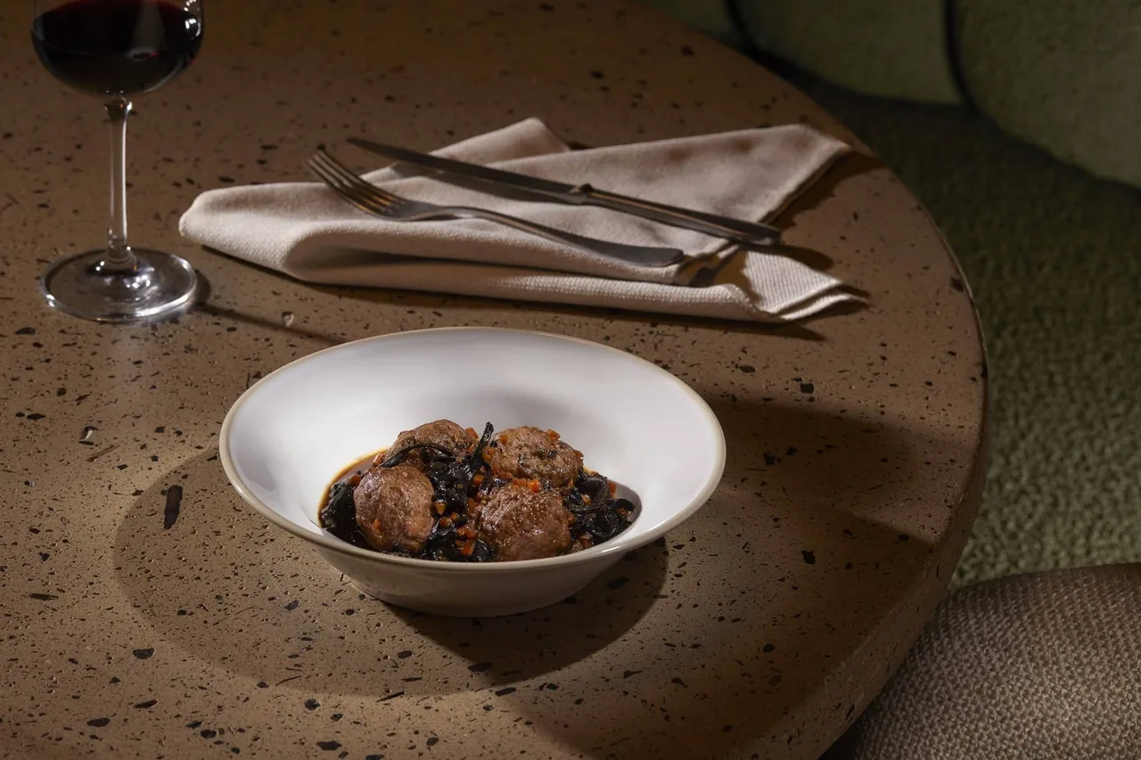 A white bowl of braised meat on a table with a glass of red wine