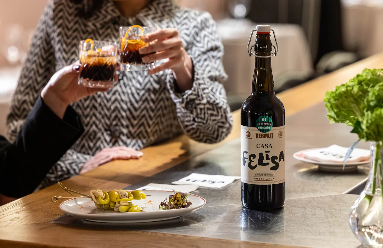 Two cut-glass tumblers of vermut with curled orange peel raised in a toast beside a bottle of Casa Felisa house vermut and a plate of gilda skewers with olives and anchovy