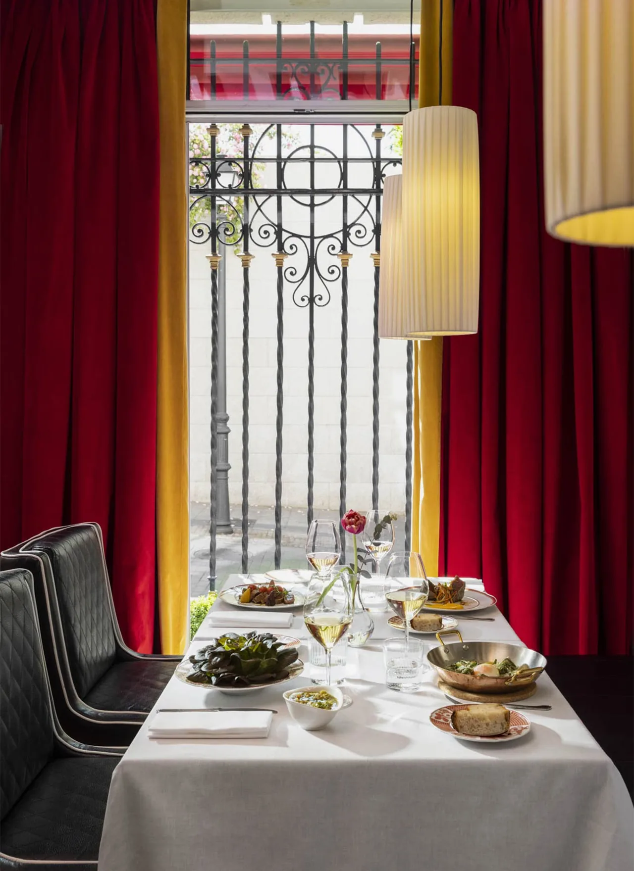 A white-clothed table by a tall window framed in red and yellow curtains, laid with several dishes including a copper pan, a plate of dark salad leaves, bread and glasses of white wine