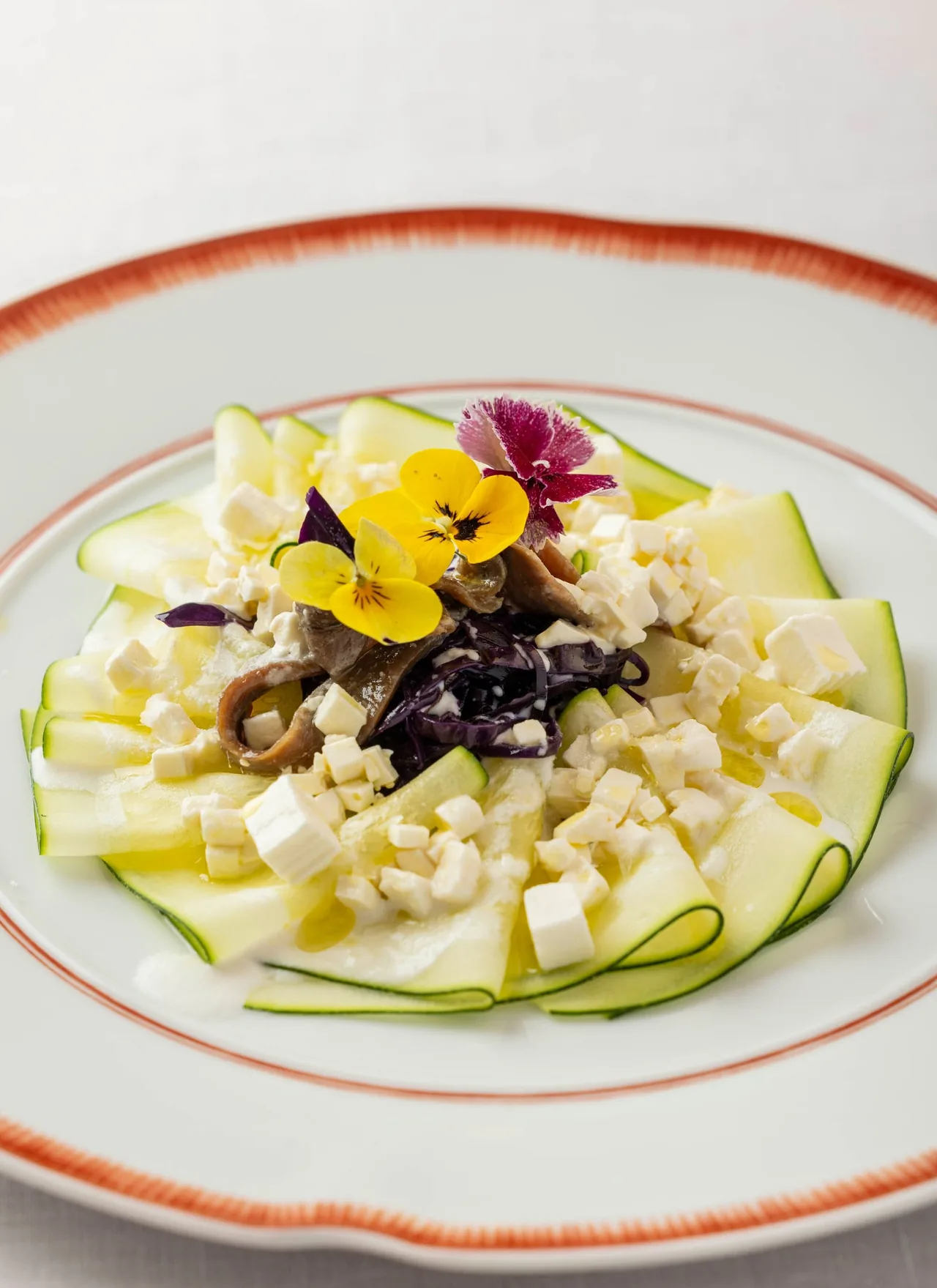 A white plate with a red rim holding sliced courgette, cheese pieces and edible flowers