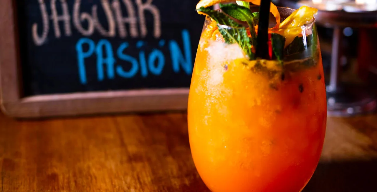 A tall glass of an orange passion-fruit cocktail with mint, dried orange and a black straw on a wooden bar top, a chalkboard behind reading 'JAGUAR PASION'
