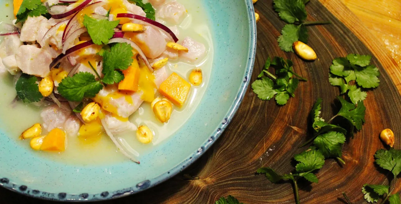A pale blue speckled bowl of ceviche, cubed white fish in leche de tigre with red onion, coriander, diced sweet potato and toasted corn kernels, on a dark wooden board with coriander scattered alongside
