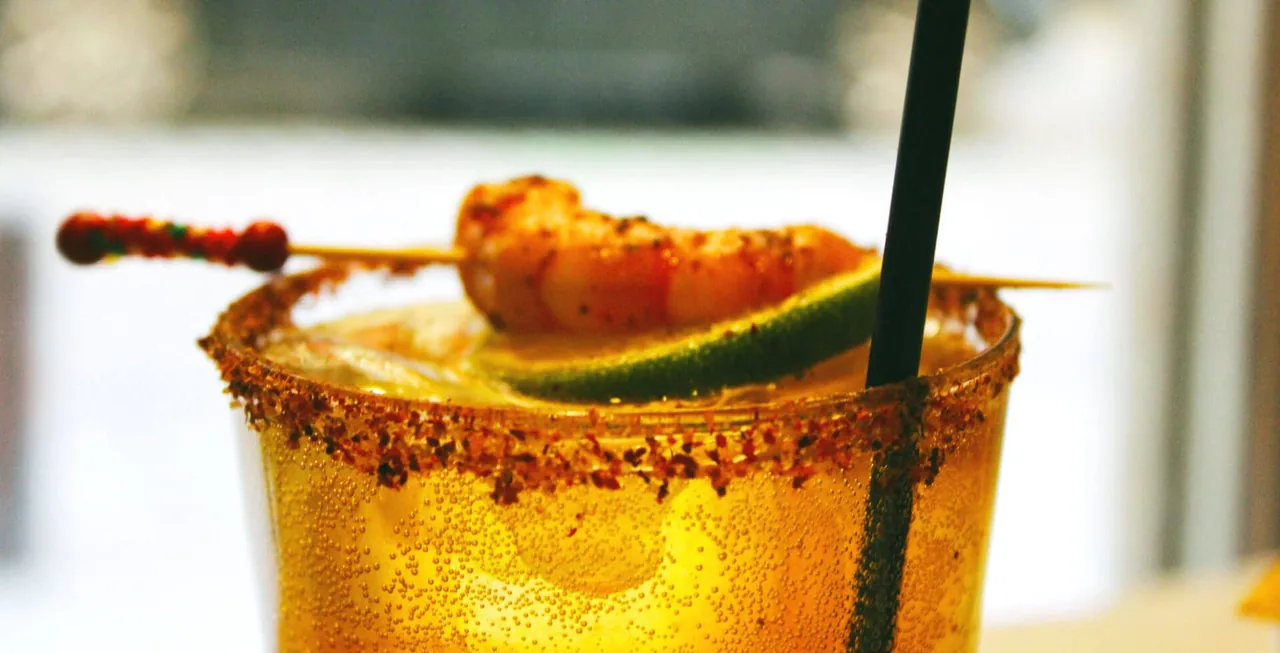 A michelada in a chilli-salt-rimmed glass, condensation on the sides, garnished with a spiced prawn on a skewer and a wedge of lime