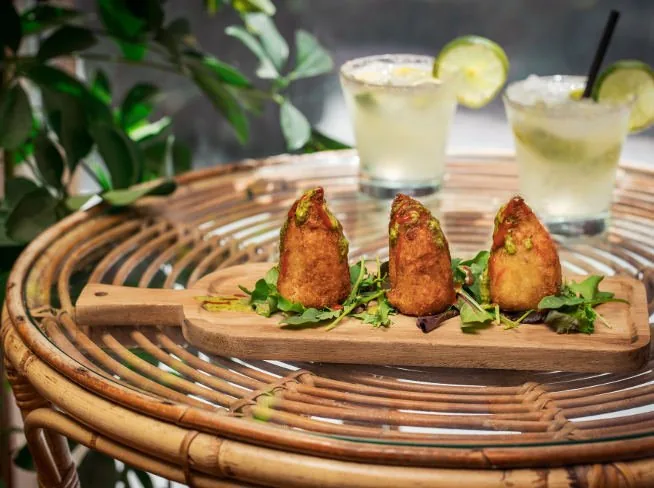 Three fried croquettes topped with pistachio crumb, standing on a wooden board over dressed leaves, two citrus cocktails on a rattan table behind