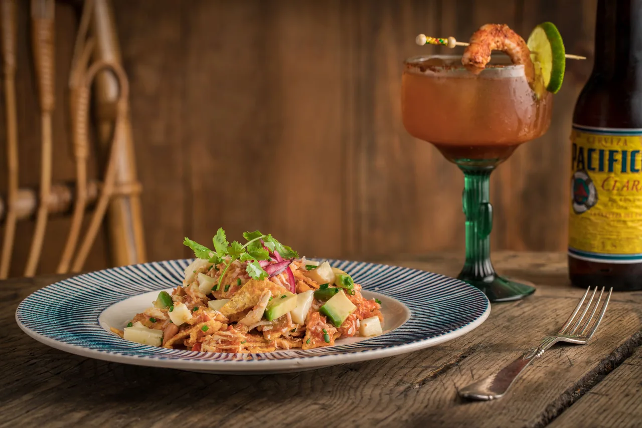 A striped plate of shredded chicken tinga with tortilla chips, avocado, jicama and coriander on a rustic wooden table, a michelada in a green-stemmed glass and a bottle of Pacifico behind