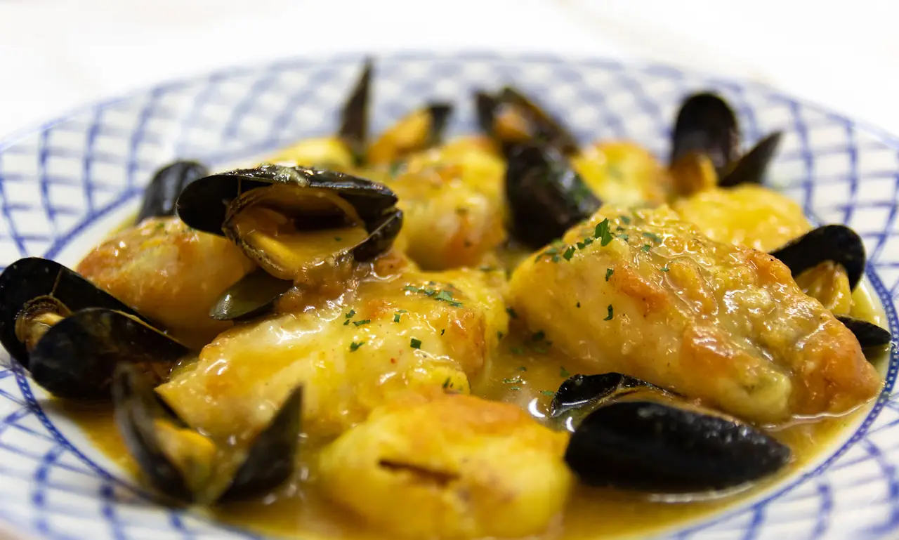 Fish fillets and mussels in a saffron-yellow sauce with potatoes, served on a blue-and-white checkered plate, at Casa Jordi