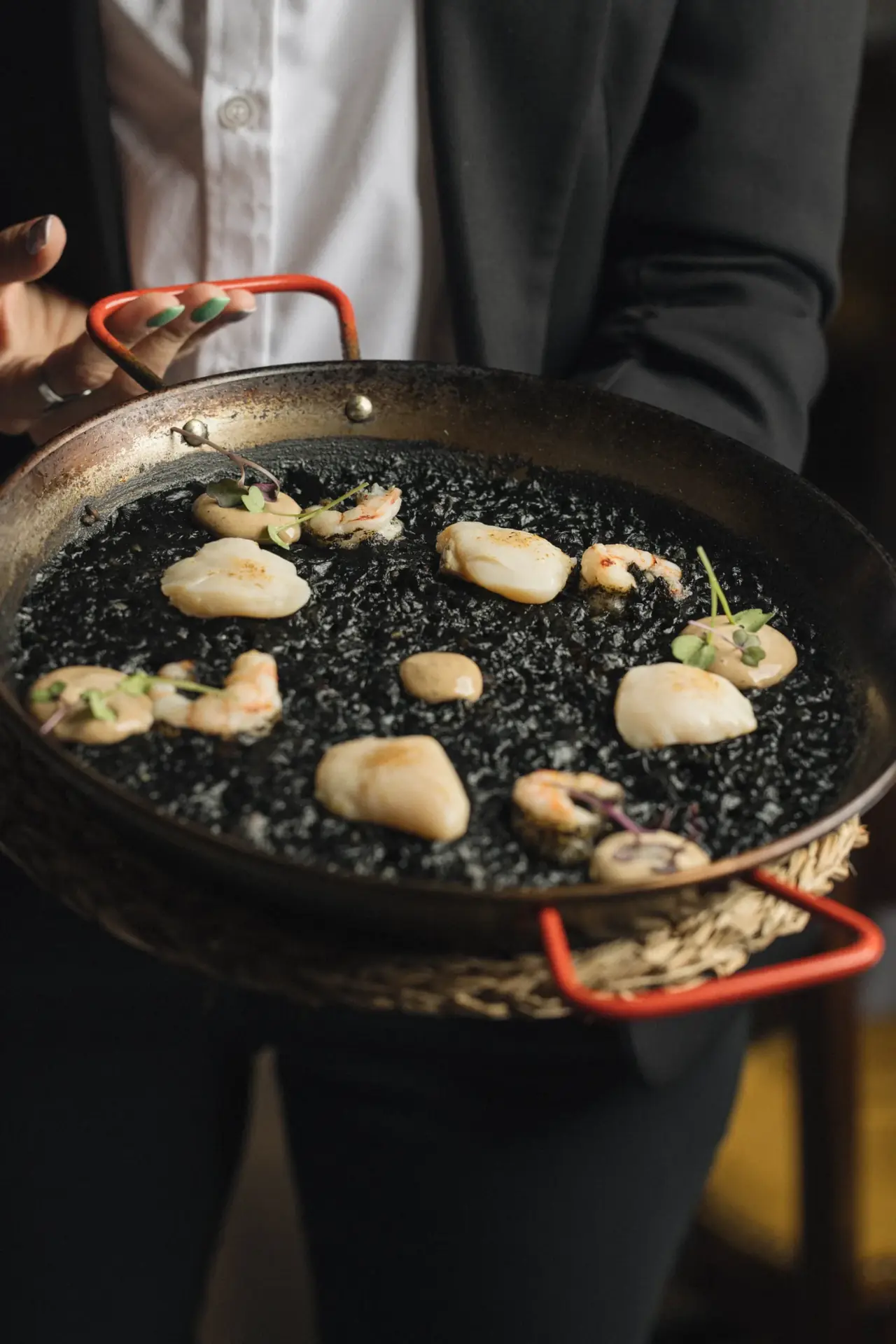 A black rice paella topped with seared scallops and shrimp in a metal pan, held by a server, at Casa Leopoldo