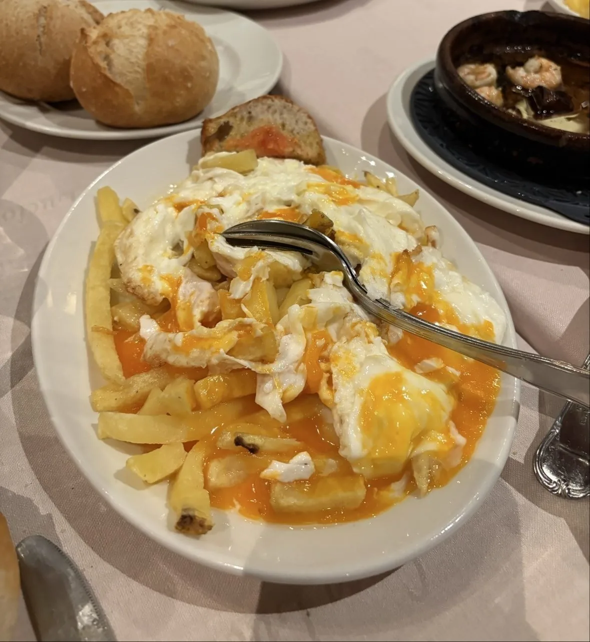 A plate of fried eggs broken over chips with a spoon, bread rolls and a clay dish of prawns behind.