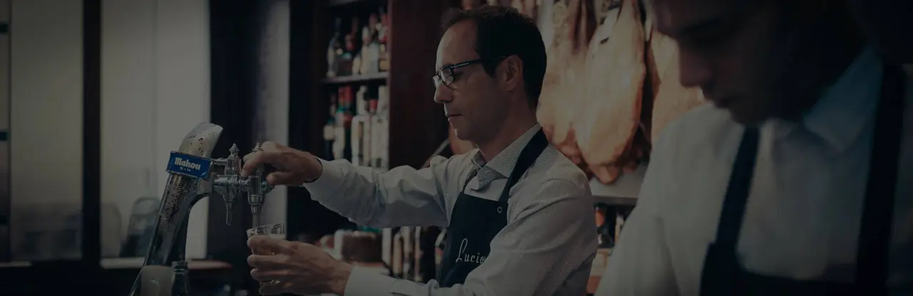 Bartender in profile pouring a beer from a Mahou tap at the bar with hams hanging behind