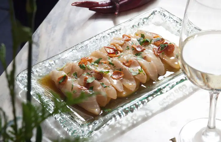 Sliced raw fish arranged on a glass plate with garlic, chili and parsley, beside a glass of white wine