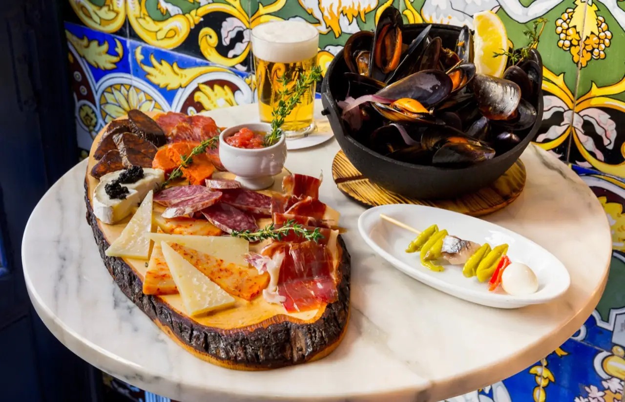 Charcuterie and cheese board with a black pot of mussels, a beer and a pickle banderilla against a tiled wall