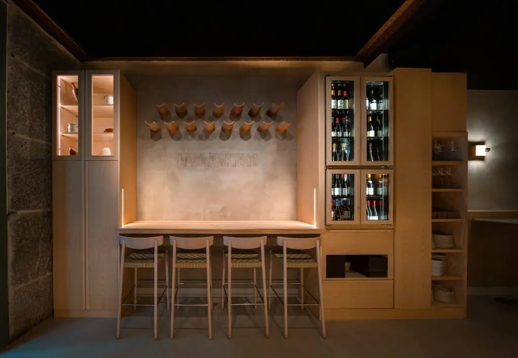 A pale wood high bar with four stools set against a plaster wall carrying the raised words CASA MORTERO, rows of wall-mounted wooden mortars above it and glass-fronted wine cabinets to one side