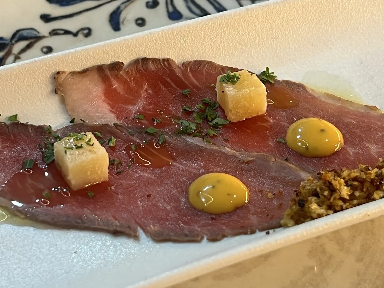 Slices of cured beef with small yellow cubes and dabs of yellow sauce on a white plate
