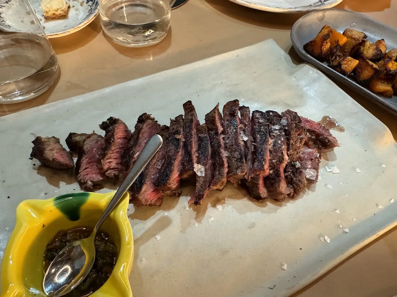 Sliced grilled steak with sea salt on a board, chimichurri and roasted potato cubes alongside